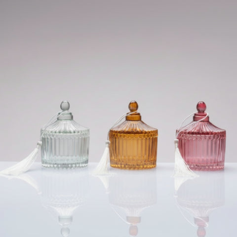 Tre scatoline portagioie in vetro lavorato con tappo e nappina bianca, mostrate nei colori grigio azzurro, ambra e rosa antico su fondo neutro